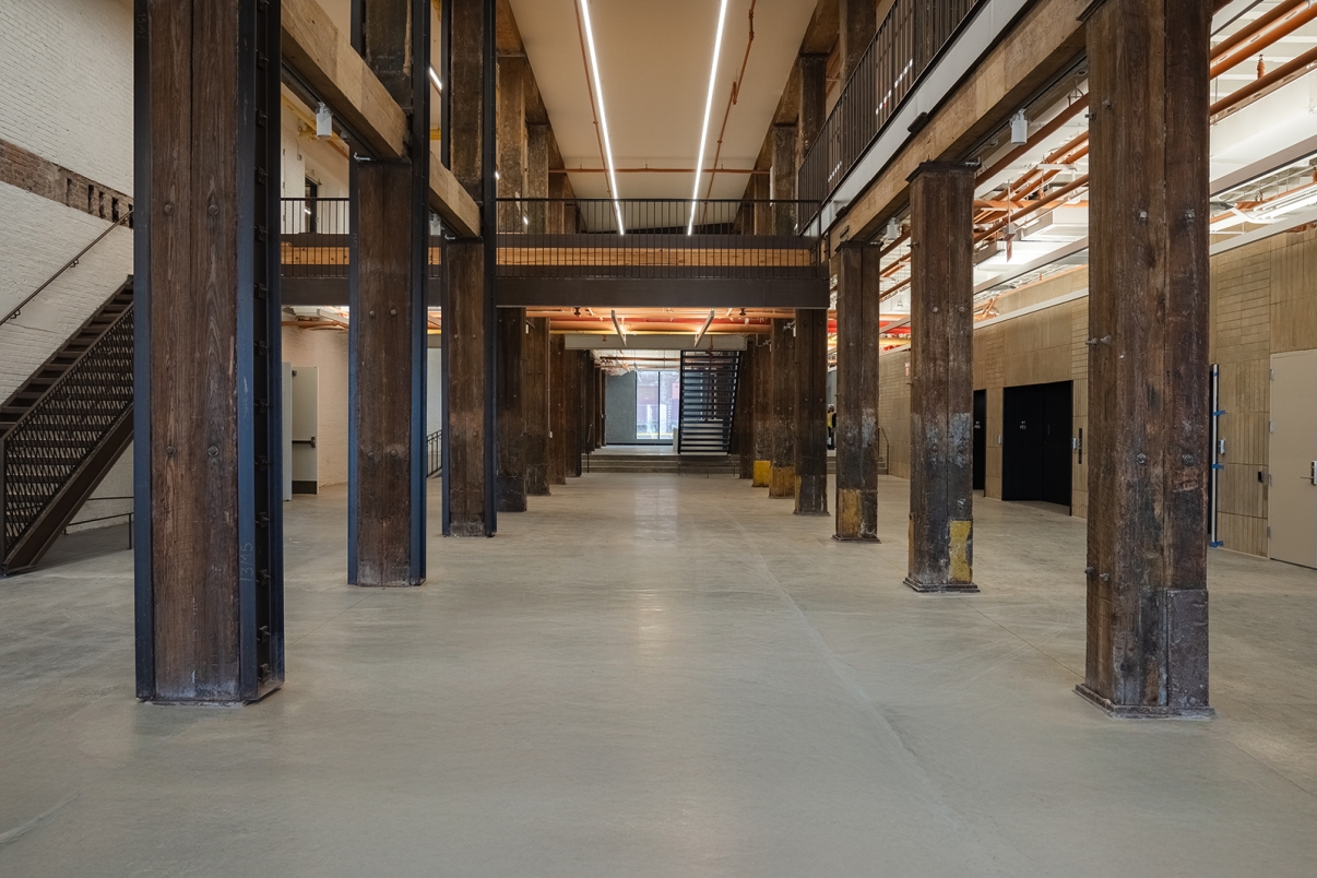 Spacious industrial interior with tall wooden columns, polished concrete floor, exposed pipes, and a central aisle leading to a staircase and glass doors at the far end.