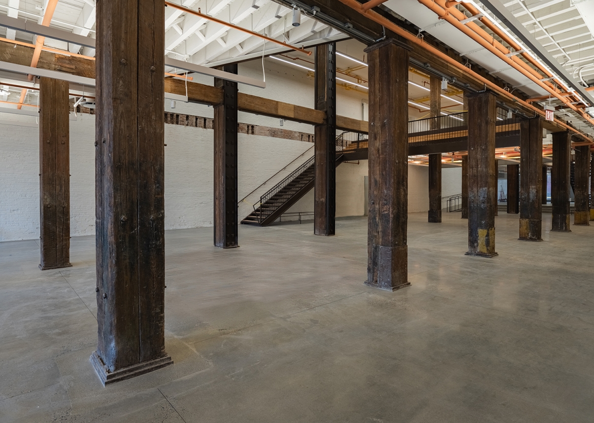 Show thumbnail preview Spacious industrial interior with large wooden beams, exposed pipes, concrete floor, white brick walls, and a metal staircase leading to a mezzanine level. The space is empty and well-lit by overhead lights.