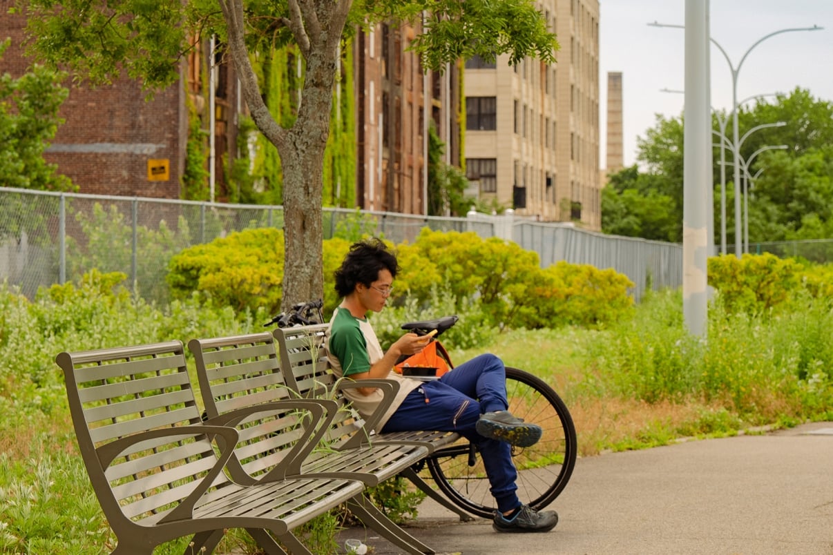 A person sits alone on a park bench, using a phone, with a bicycle propped nearby. Lush greenery and a large tree surround the area, and an industrial building is visible in the background.
