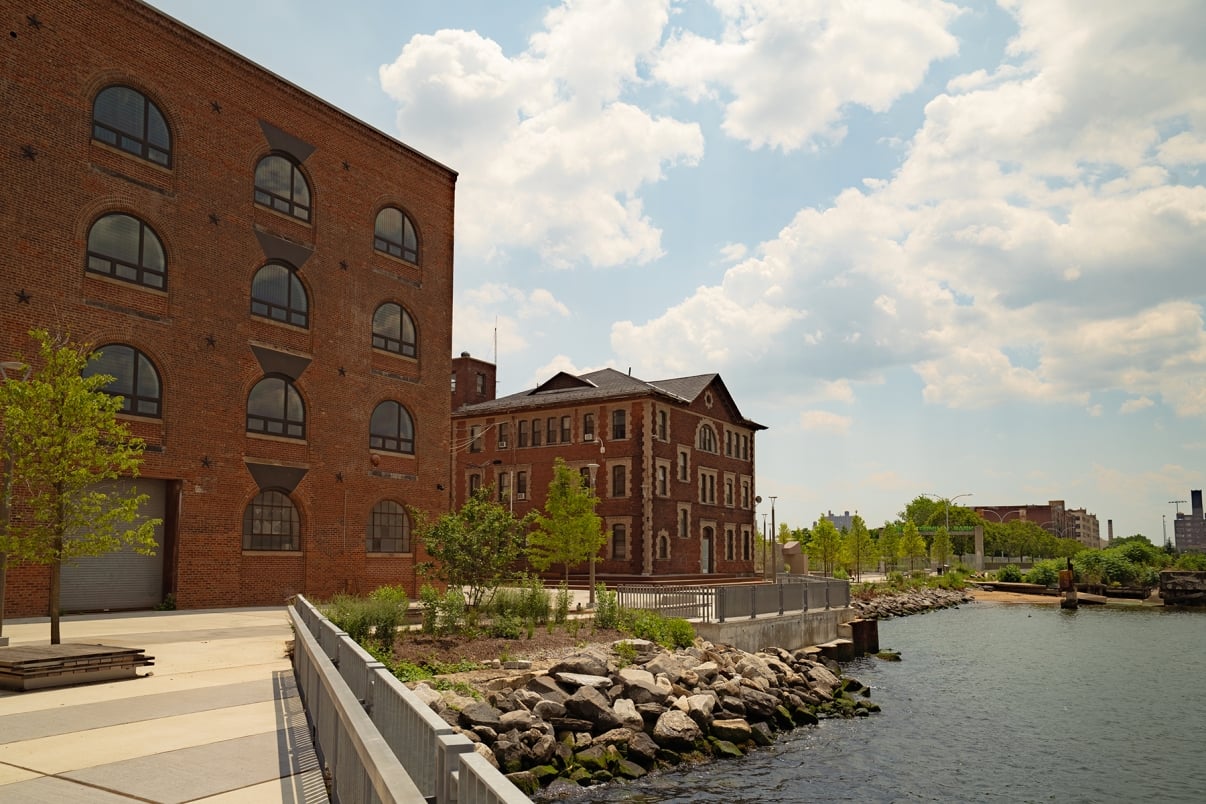 Red brick industrial buildings sit beside a river, surrounded by trees and rocks along the water’s edge. The sky is partly cloudy, and the area appears calm and well-maintained.