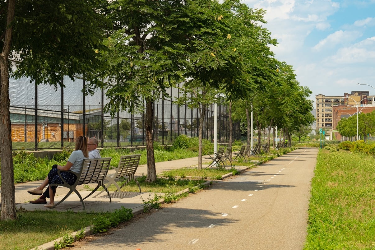 Two people sit on a bench under the shade of trees along a pathway, with empty benches and a fenced sports area to the left and buildings visible in the background on a sunny day.