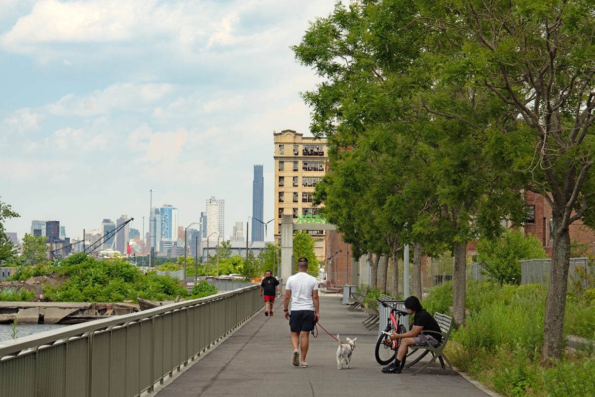 A man walks a dog on a riverside path lined with trees; another man jogs, and a third sits on a bench by a bicycle. City skyline and buildings are visible in the background under a partly cloudy sky.