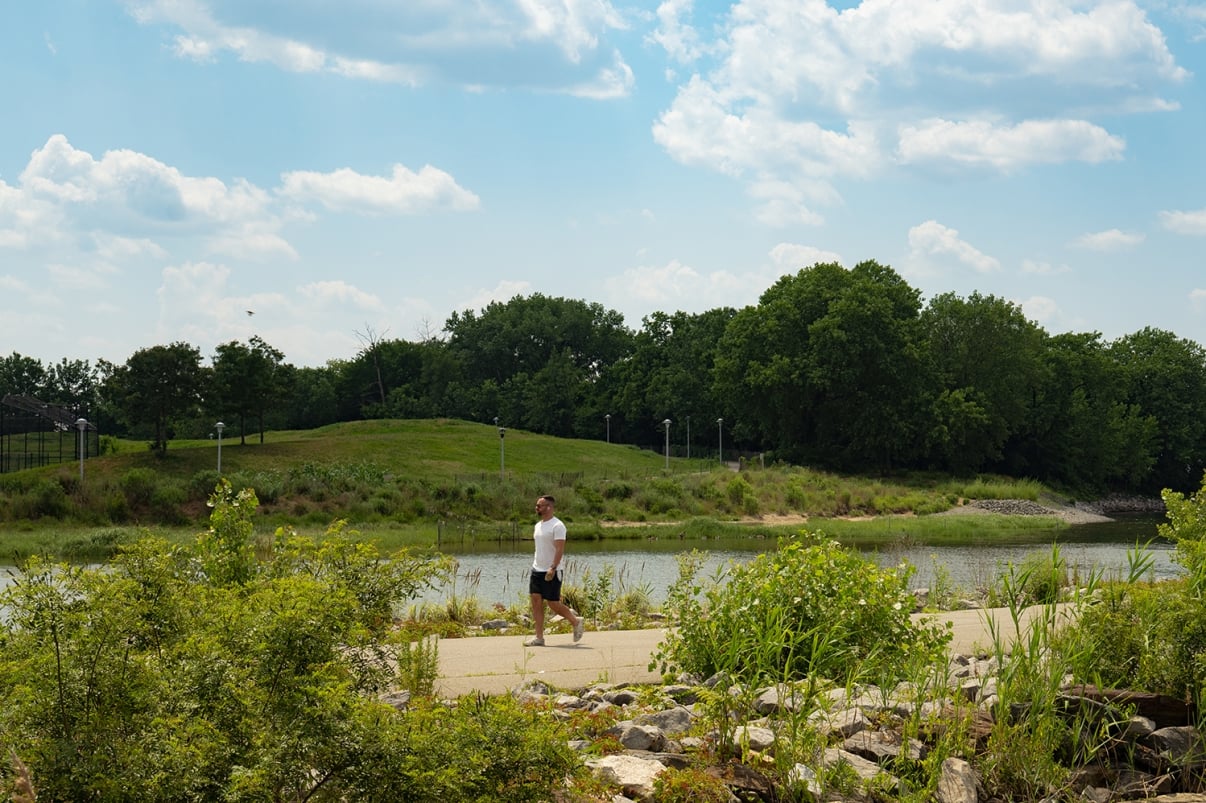 A man in a white shirt and black shorts walks on a paved path beside a river, surrounded by green bushes and trees under a partly cloudy sky.