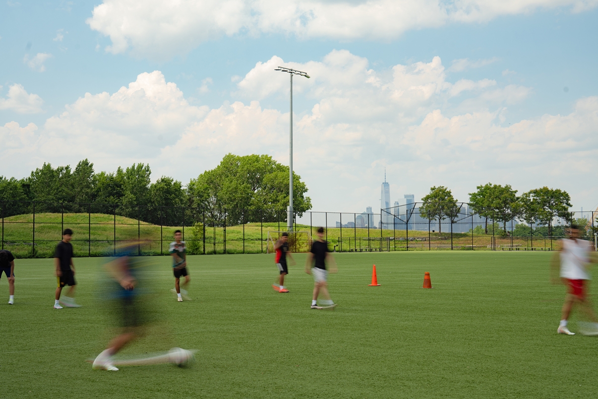 A group of people play soccer on a green field with orange cones, some in motion blur. Trees and a city skyline with tall buildings are visible in the background under a partly cloudy sky.