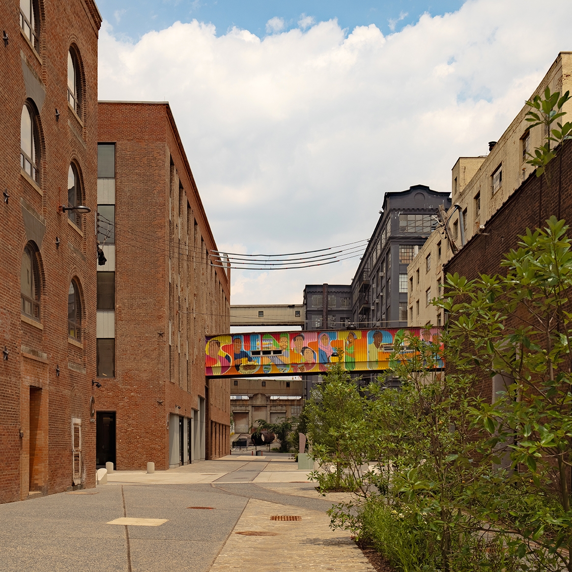 A quiet urban alley lined with brick and concrete buildings, featuring a colorful mural-covered pedestrian bridge connecting two structures, with greenery on the right and a partly cloudy sky above.