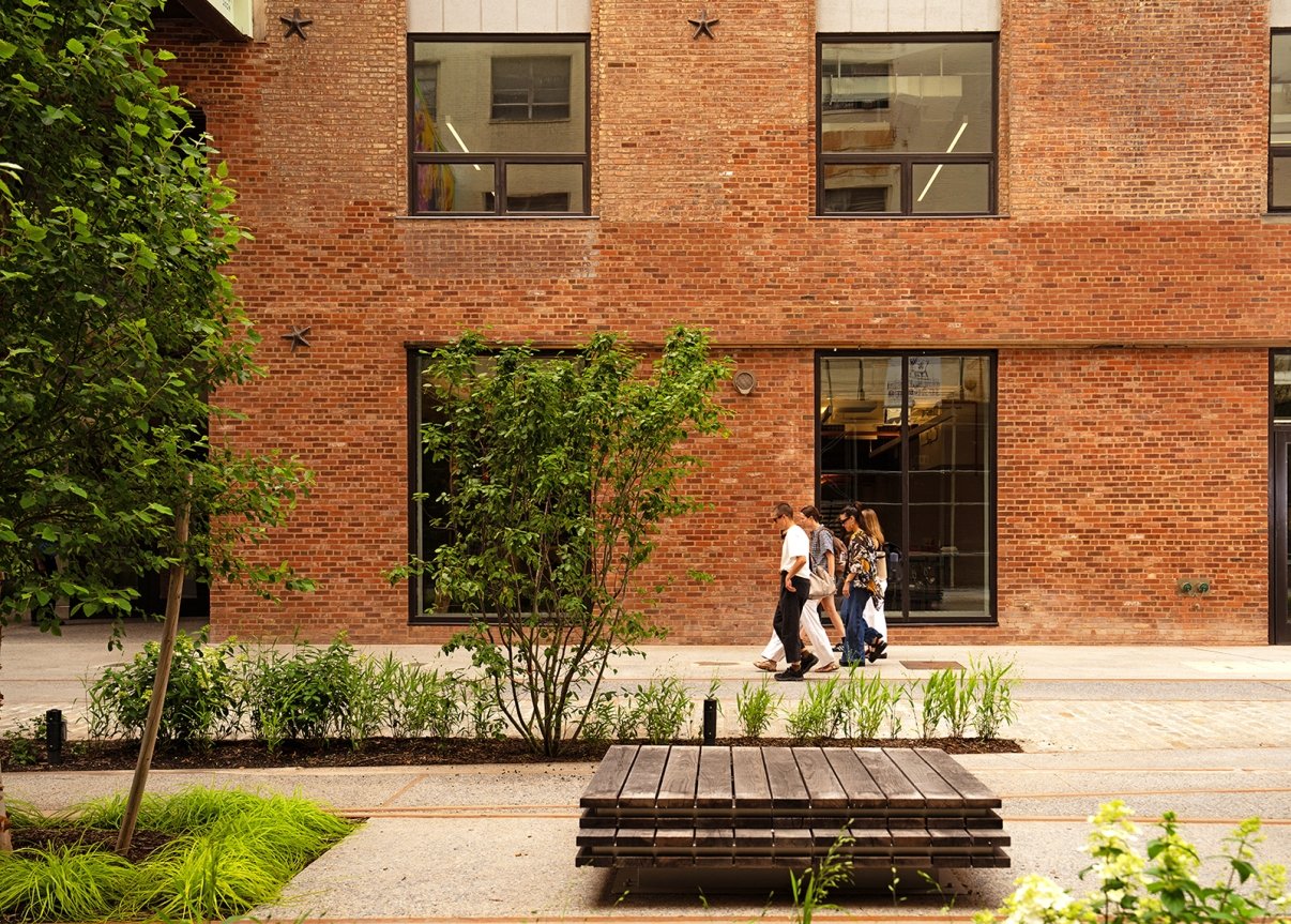 Four people walk along a sidewalk next to a brick building with large windows, surrounded by greenery and trees. A wooden bench sits in the foreground of the urban park setting.
