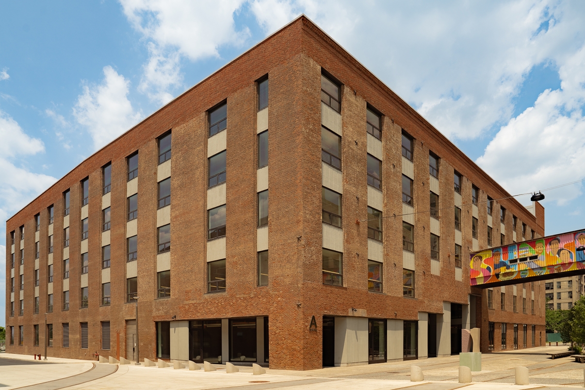 A large, rectangular brick building with tall windows, flat roof, and a colorful mural on a skybridge attached to the side, under a blue sky with scattered clouds.