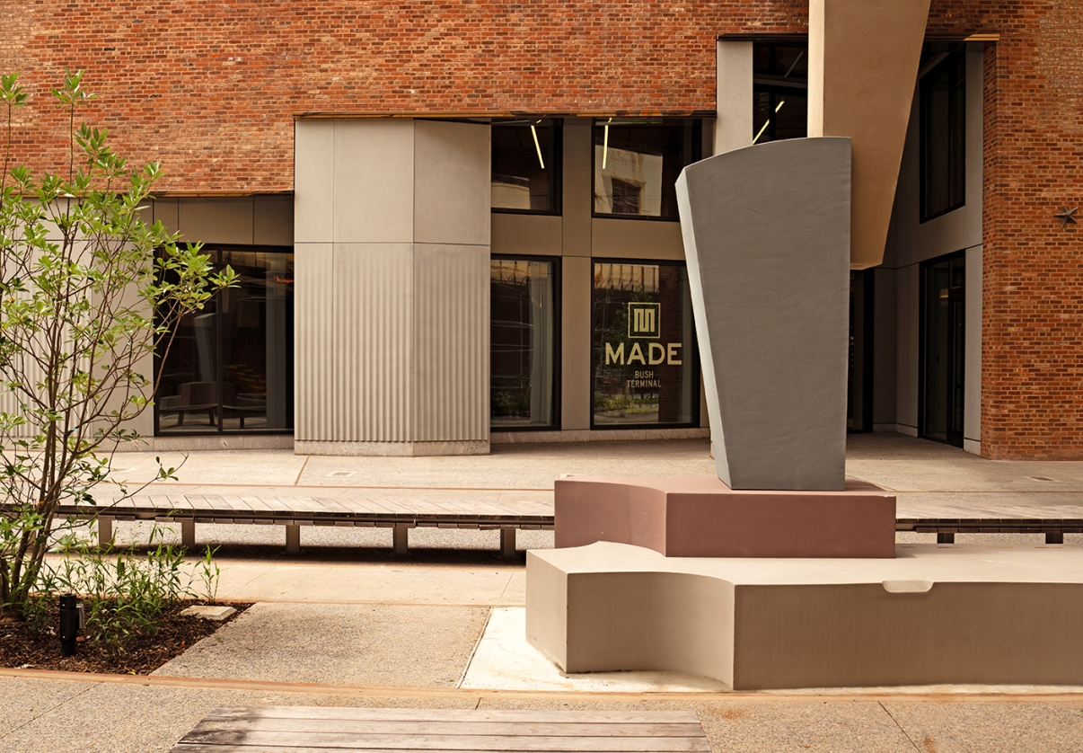 A modern outdoor courtyard with abstract concrete benches and a geometric sculpture, set against a brick building with large windows. One window displays the words MADE in bold letters.
