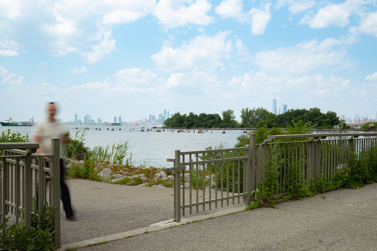 A blurred person walks past a metal fence near a waterfront with distant city skyline and trees under a partly cloudy sky.