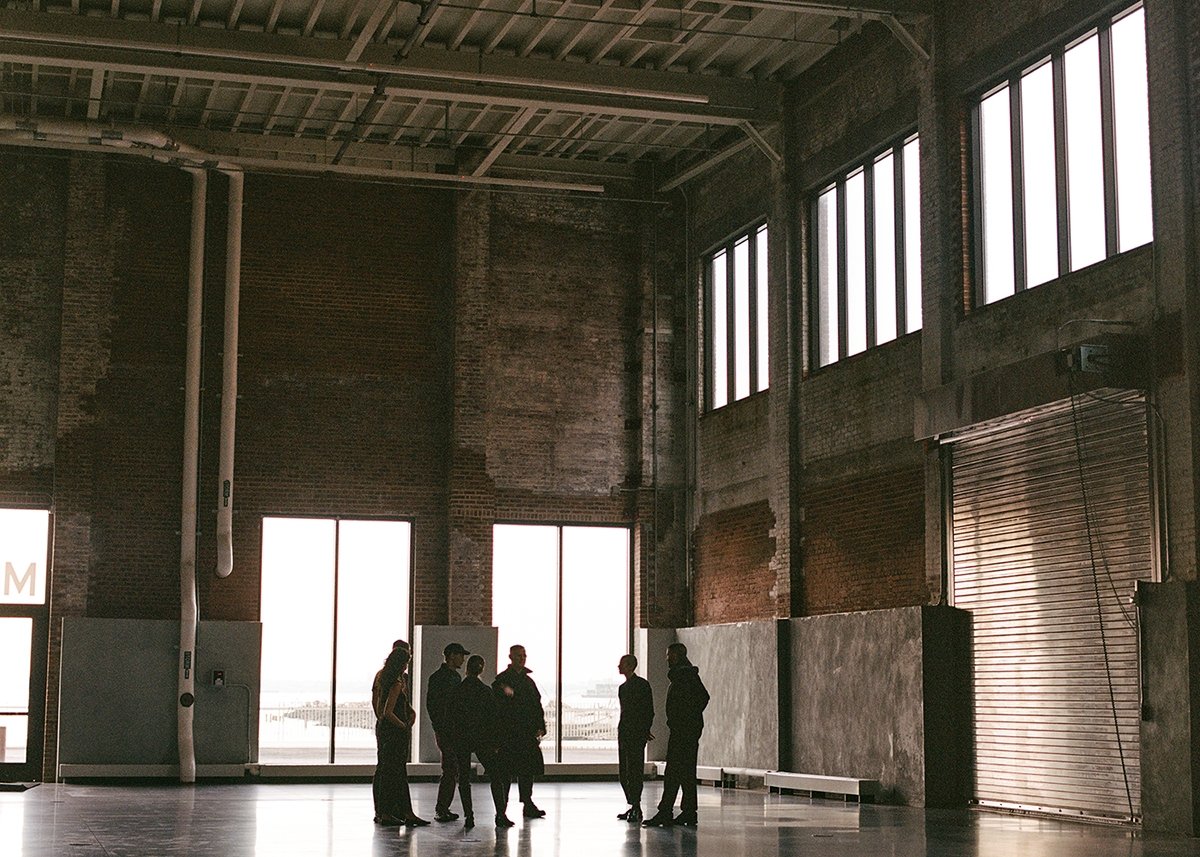 A group of people stand and talk inside a large, industrial warehouse with tall windows, exposed brick walls, and a high ceiling. Light streams in through the windows, casting shadows on the floor.